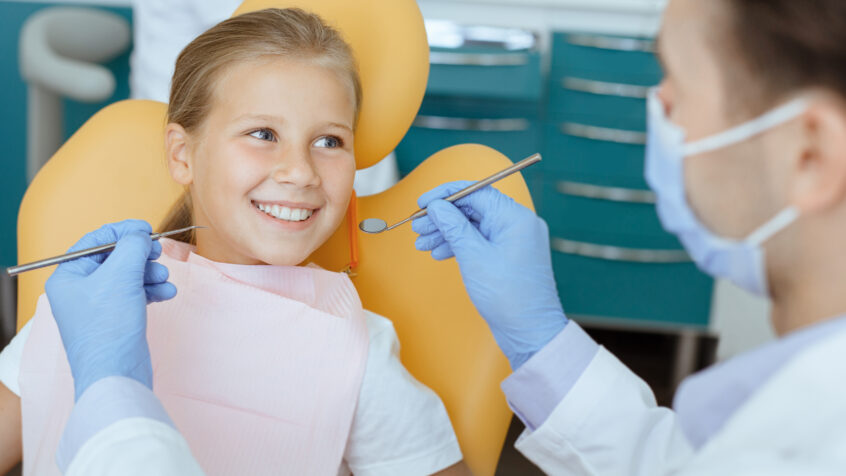 Modern painless dental treatment for children. Doctor in protective mask, white coat and rubber gloves with tools examines small smiling kid in chair in interior of modern clinic